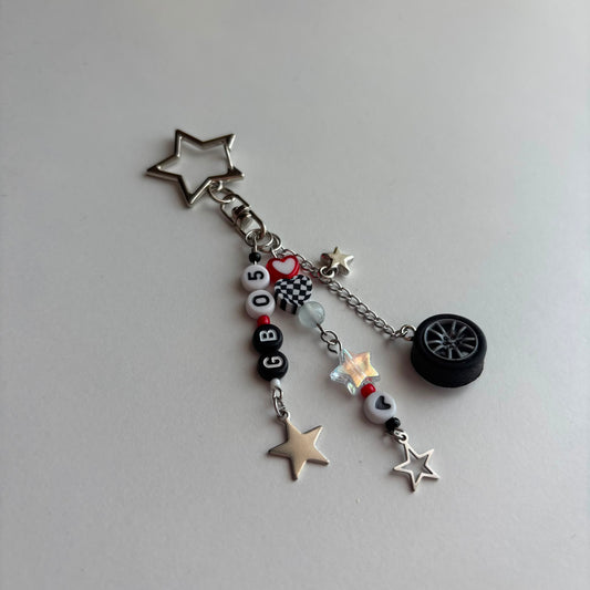 A silver-toned keychain charm featuring a star-shaped clasp and multiple dangling charms, including a miniature black tire, silver stars, a checkered flag bead, and white beads spelling out 'GB05'.