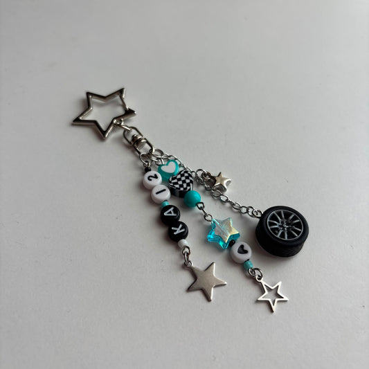 A silver-tone keychain charm featuring a star-shaped keyring, a chain with beads, in white, black, and turquoise, a checkered pattern bead, a turquoise star, and a miniature black tire charm, all on a light gray background.
