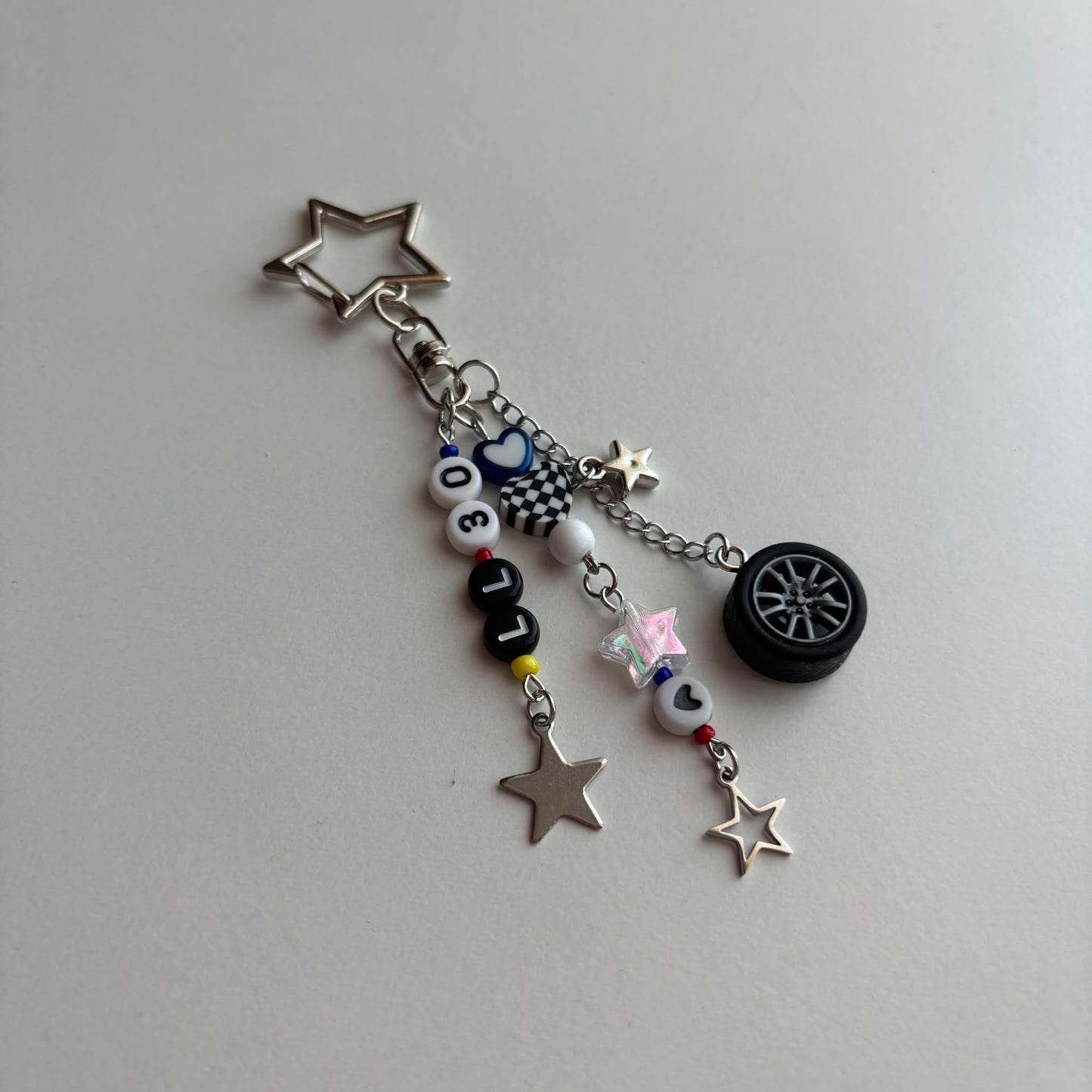 A silver metal keychain charm featuring a star-shaped ring, a checkered flag charm, a miniature black tire, and white beads spelling out 'LL30' with small accent beads.