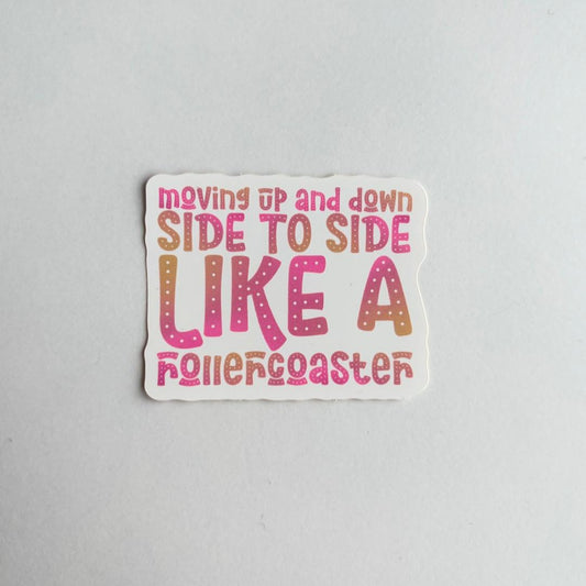 A die-cut sticker with the phrase 'moving up and down SIDE TO SIDE LIKE A rollercoaster' in pink, orange, and brown gradient lettering on a white background, photographed on a light gray surface.