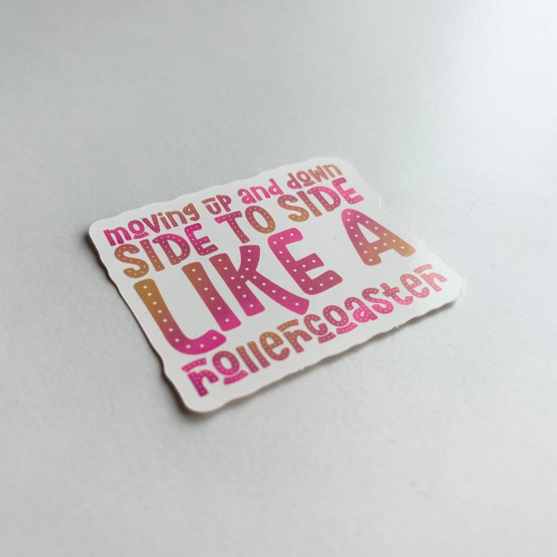 A die-cut sticker with a white background and colorful text that reads 'moving up and down SIDE TO SIDE LIKE A rollercoaster' in pink, orange, and brown gradient lettering, shown on a light gray surface.