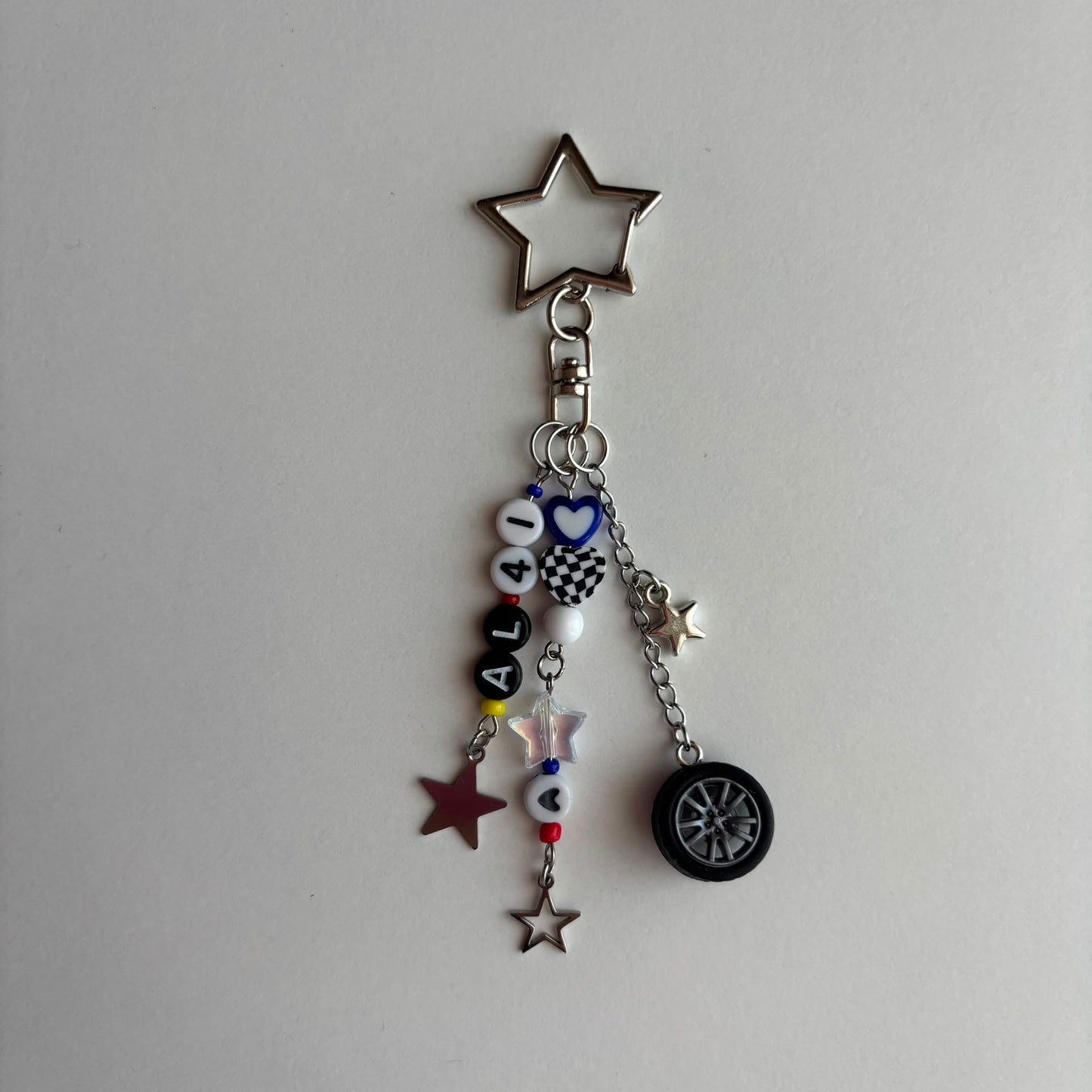 A silver metal keychain charm featuring a star-shaped top, a clasp, and dangling charms including white beads spelling 'AL41', a checkered heart, a small tire charm, and star charms on a plain light background.