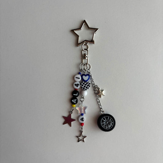 A silver metal keychain charm featuring a star-shaped top, a clasp, and dangling charms including white beads spelling 'AL41', a checkered heart, a small tire charm, and star charms on a plain light background.