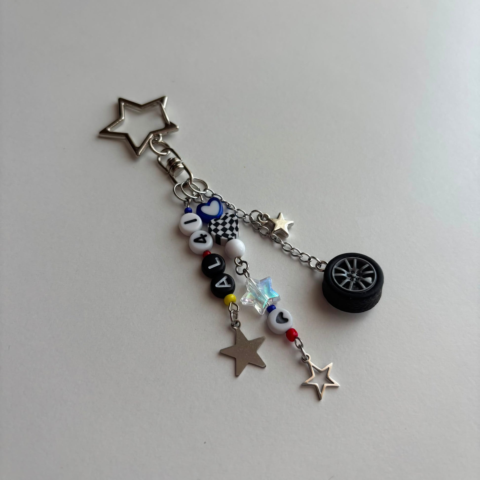 A silver-toned keychain charm featuring a star-shaped clasp, a small tire charm, and dangling beads and star charms on a plain light gray background.