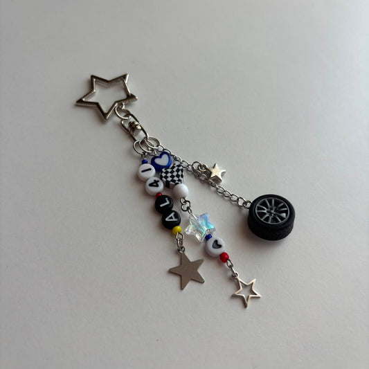 A silver-toned keychain charm featuring a star-shaped clasp, a small tire charm, and dangling beads and star charms on a plain light gray background.