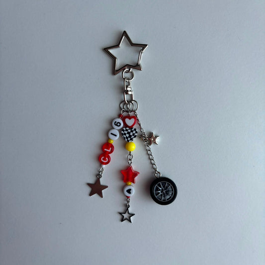 A silver metal keychain charm featuring a large star-shaped top, a clasp, and dangling charms including red and white beads spelling 'CL16', a checkered pattern, a small red star, a black tire charm, and several star charms, displayed on a light gray background.