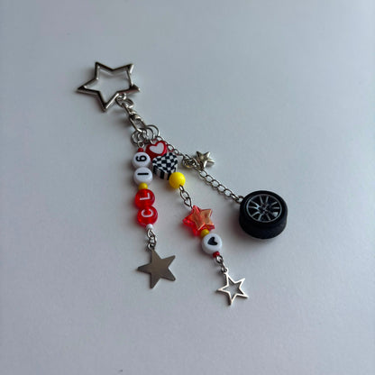 A silver-toned metal charm keychain featuring a star-shaped keyring, a short chain, and multiple dangling charms including a black car wheel, silver stars, a checkered flag, red and white beads, and a small yellow bead, photographed on a plain light gray surface.