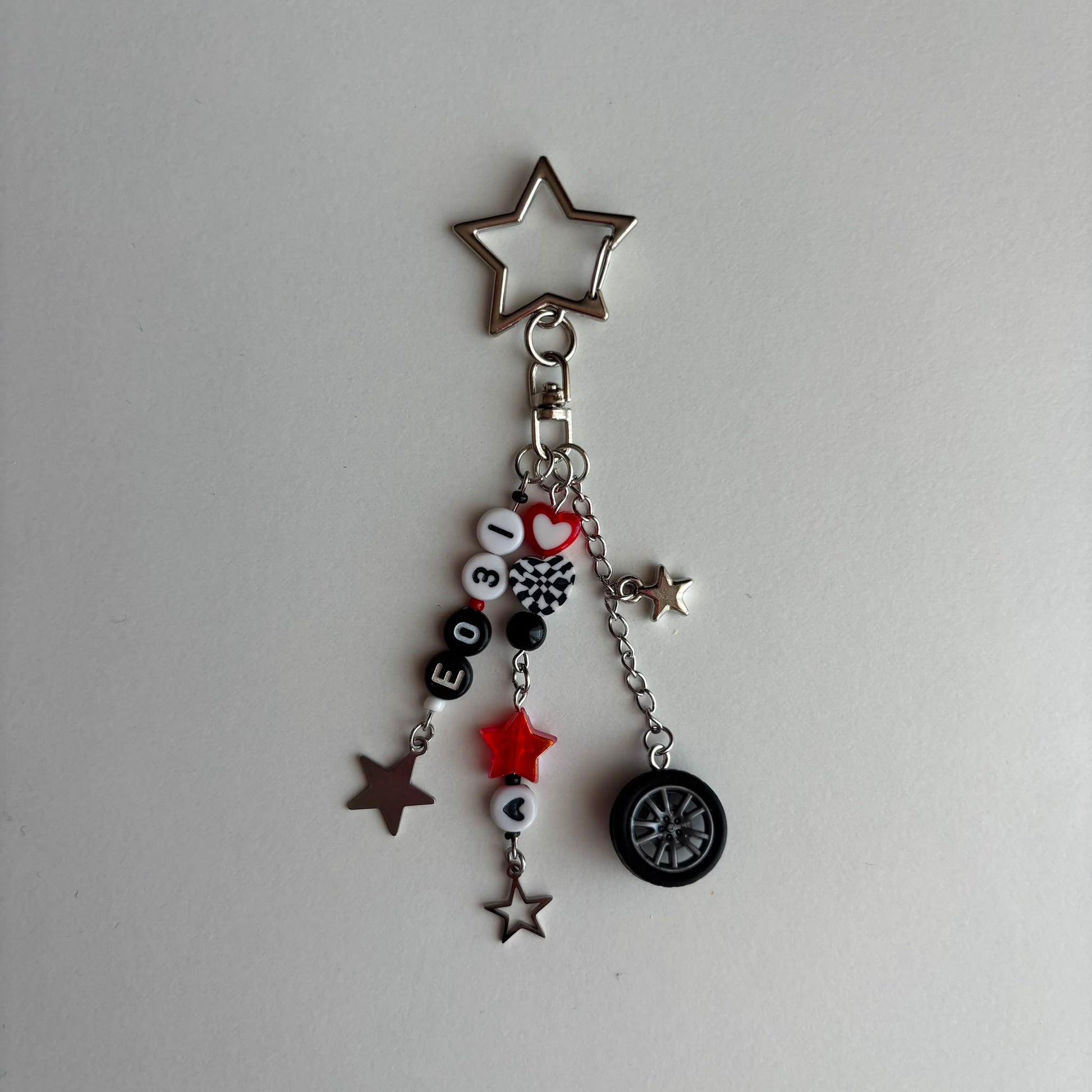 A silver-toned metal keychain charm featuring a star-shaped clasp and dangling charms, including a small black tire, beads spelling 'EO31', a red heart, and several star charms, shown on a plain light gray background.