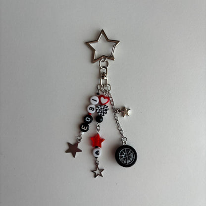 A silver-toned metal keychain charm featuring a star-shaped clasp and dangling charms, including a small black tire, beads spelling 'EO31', a red heart, and several star charms, shown on a plain light gray background.