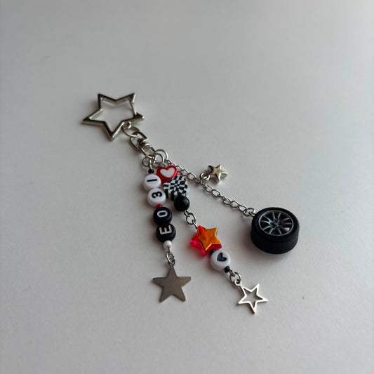 A silver-toned keychain charm featuring a star-shaped metal clasp, a miniature black tire charm, and multiple dangling charms including silver stars, black and white beads spelling 'EO31', a checkered pattern bead, a red heart, and a small orange star, all photographed on a plain light gray surface.