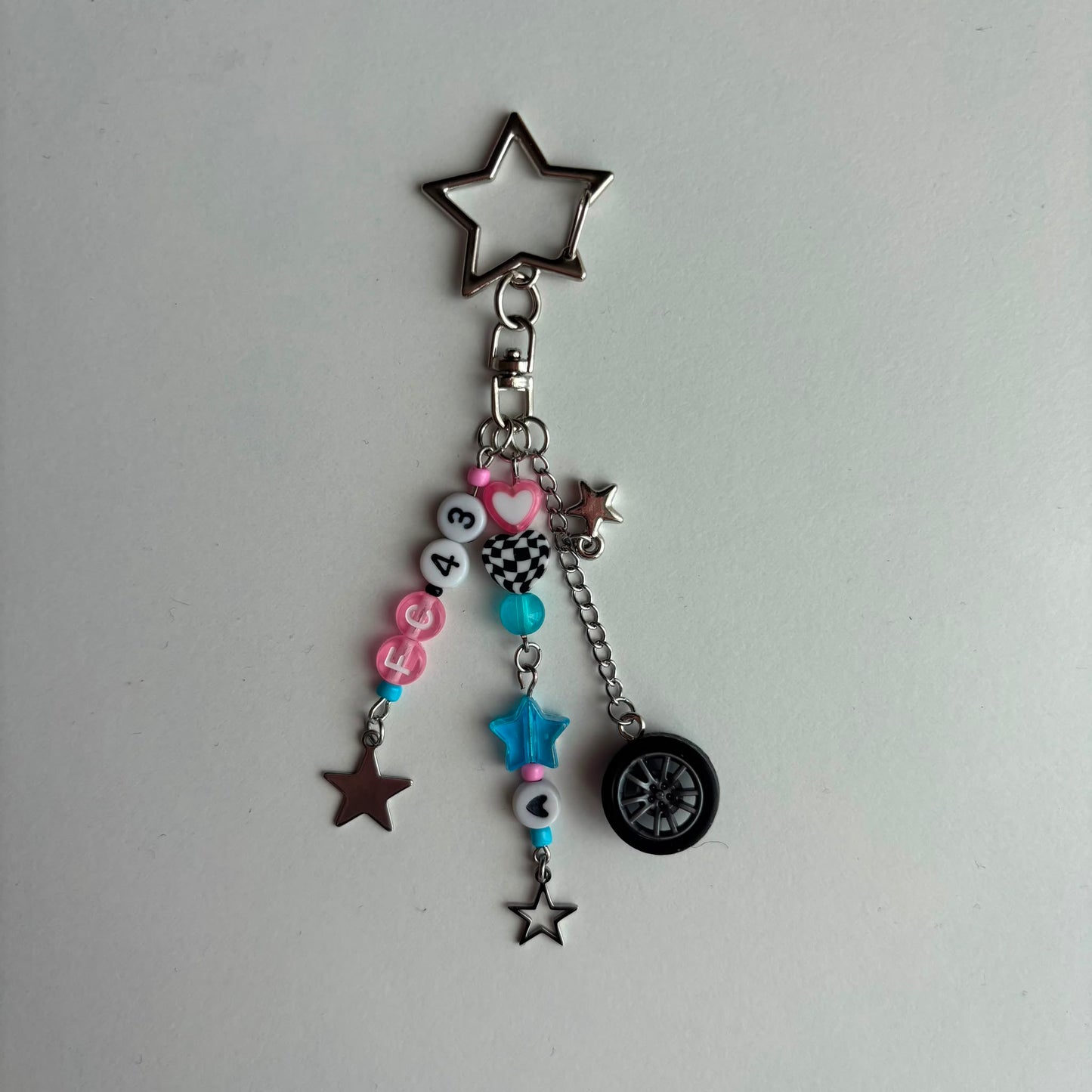 A silver metal keychain charm featuring a star-shaped ring, a beaded chain with pink and blue beads, a checkered bucket charm, and a small black tire charm, displayed on a plain light gray surface.