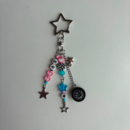 A silver metal keychain charm featuring a star-shaped ring, a beaded chain with pink and blue beads, a checkered bucket charm, and a small black tire charm, displayed on a plain light gray surface.