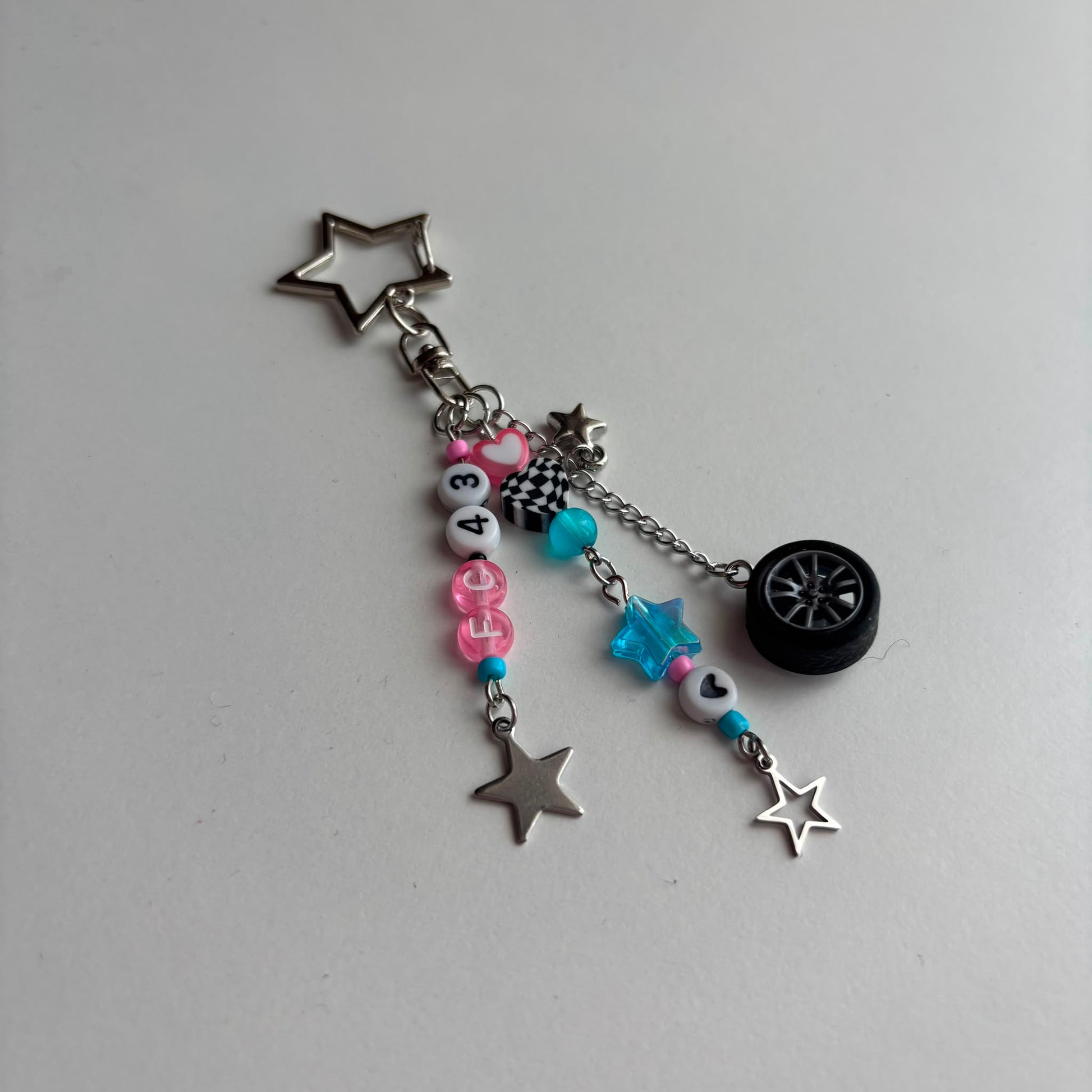 A silver-toned keychain with automotive-themed charms, including a star-shaped charm, a miniature tire, a checkered flag, and pink and blue beads on a light gray background.