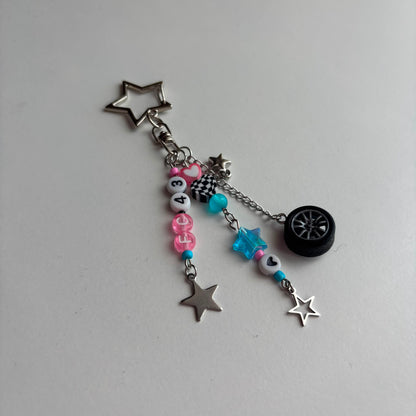 A silver-toned keychain with automotive-themed charms, including a star-shaped charm, a miniature tire, a checkered flag, and pink and blue beads on a light gray background.