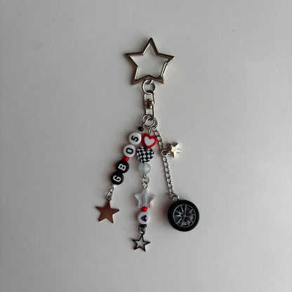 A silver-toned keychain charm featuring a star-shaped top, a clasp, and dangling charms including letter beads spelling 'GB05', a checkered heart, a star, a tire, and small star charms on a plain light background.
