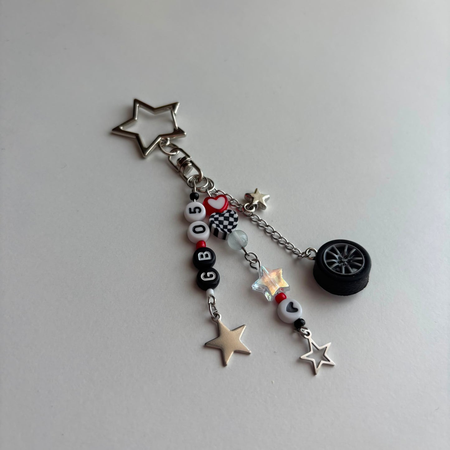 A silver-toned keychain charm featuring a star-shaped clasp and multiple dangling charms, including a miniature black tire, silver stars, a checkered flag bead, and white beads spelling out 'GB05'.