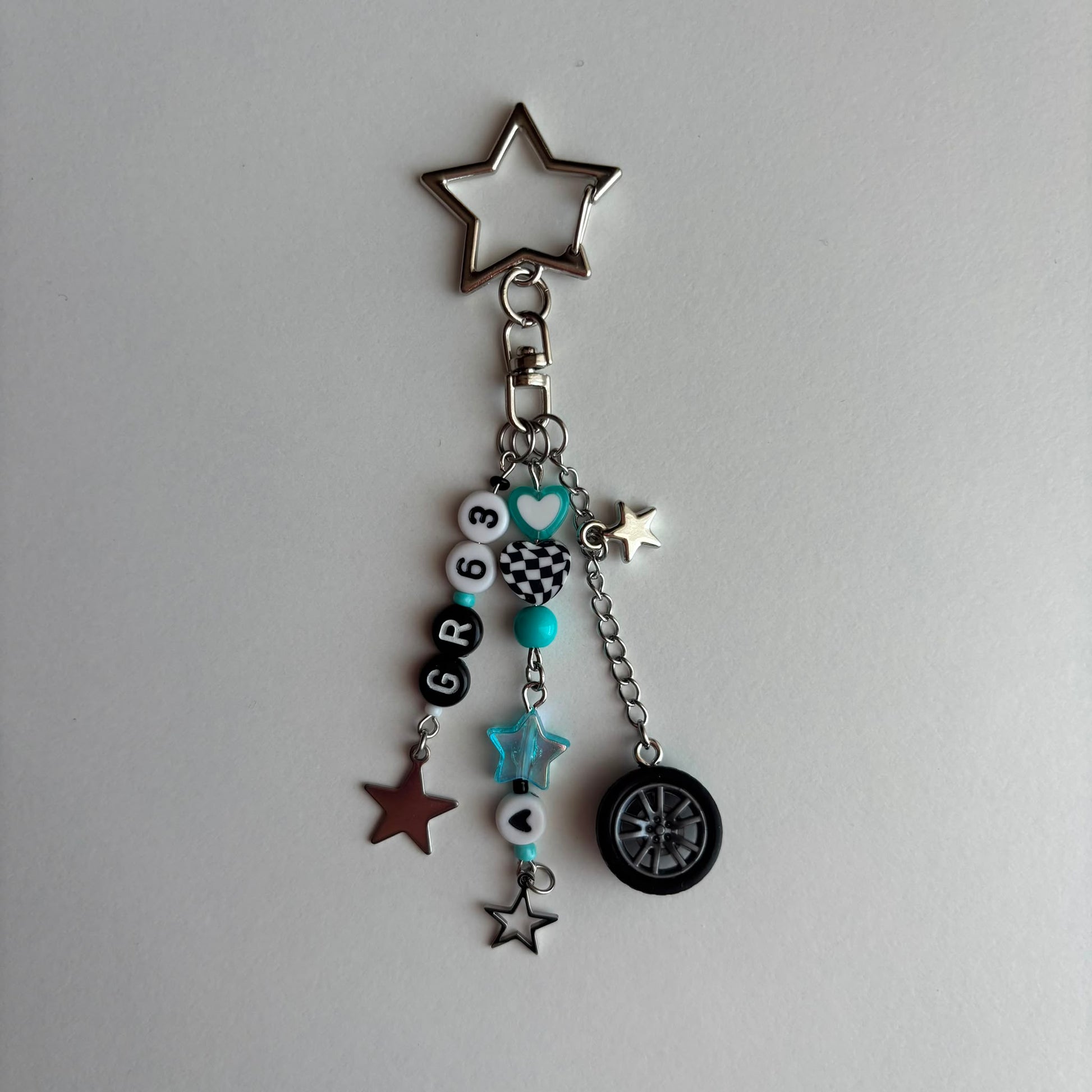 A silver metal keychain charm featuring a star-shaped ring at the top, a clasp, and multiple dangling charms including black and white beads with numbers and checkered patterns, turquoise accents, star charms and a tire-shaped charm, all displayed on a light gray background.