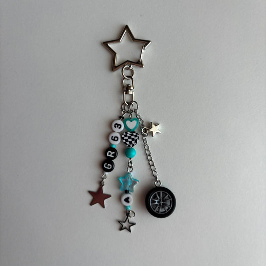A silver metal keychain charm featuring a star-shaped ring at the top, a clasp, and multiple dangling charms including black and white beads with numbers and checkered patterns, turquoise accents, star charms and a tire-shaped charm, all displayed on a light gray background.