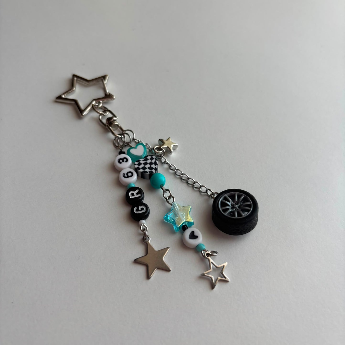 A silver and black car-themed charm keychain featuring a star-shaped metal ring, a small black tire charm, turquoise beads, checkered details, and letter beads spelling 'GR63', displayed on a light gray background.