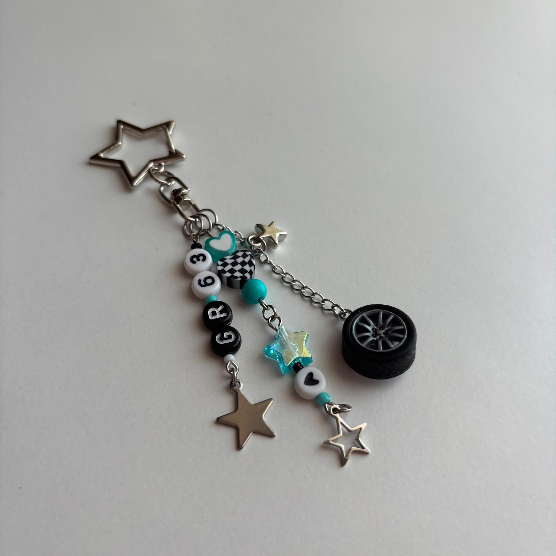A silver and black car-themed charm keychain featuring a star-shaped metal ring, a small black tire charm, turquoise beads, checkered details, and letter beads spelling 'GR63', displayed on a light gray background.