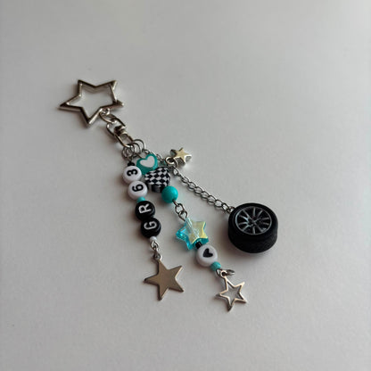 A silver and black car-themed charm keychain featuring a star-shaped metal ring, a small black tire charm, turquoise beads, checkered details, and letter beads spelling 'GR63', displayed on a light gray background.