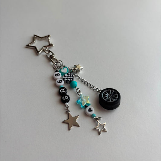 A silver and black car-themed charm keychain featuring a star-shaped metal ring, a small black tire charm, turquoise beads, checkered details, and letter beads spelling 'GR63', displayed on a light gray background.