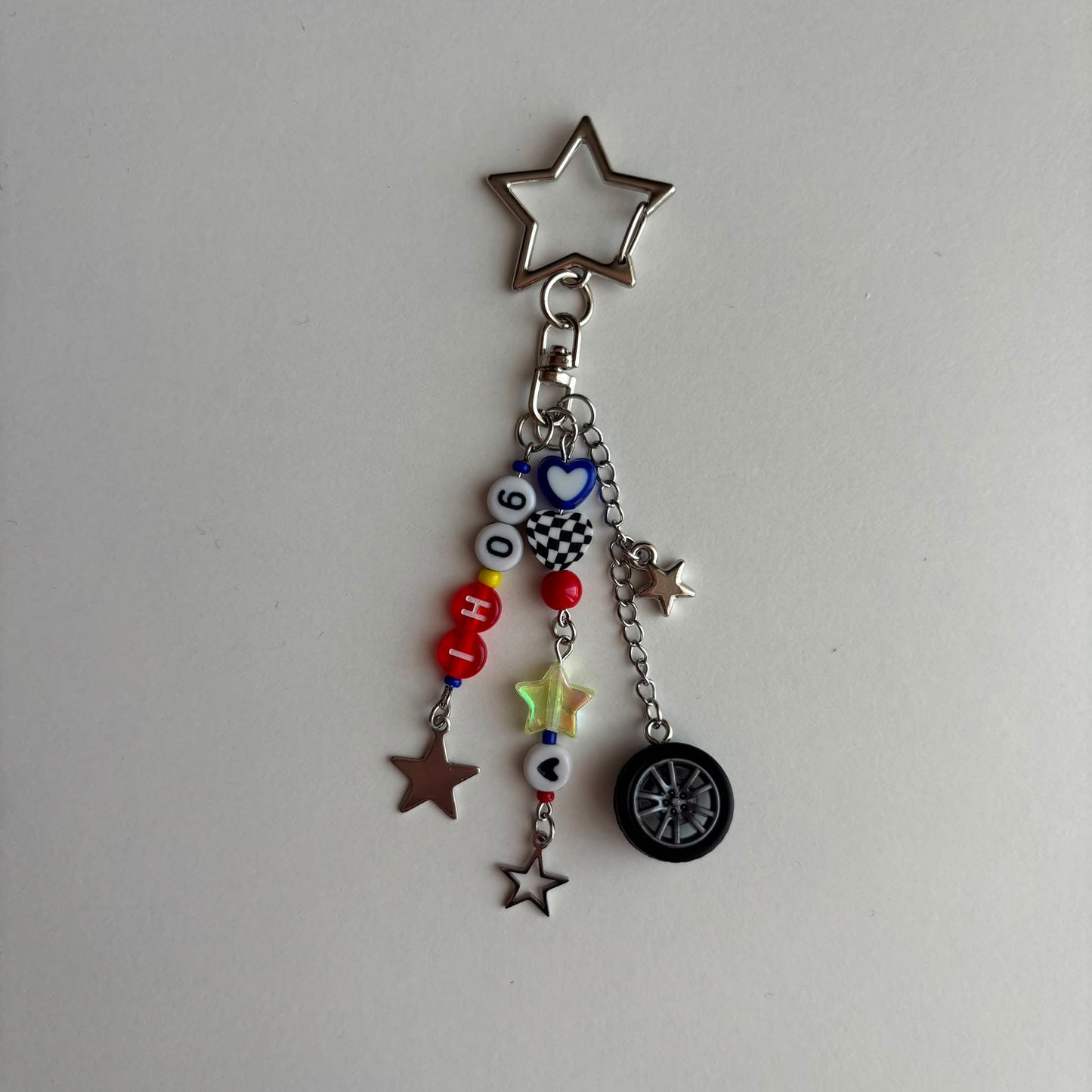 A silver metal keychain charm featuring a star-shaped clasp and dangling charms, including a small tire, star cutouts, checkered pattern, and colorful beads spelling out 'IH06'.