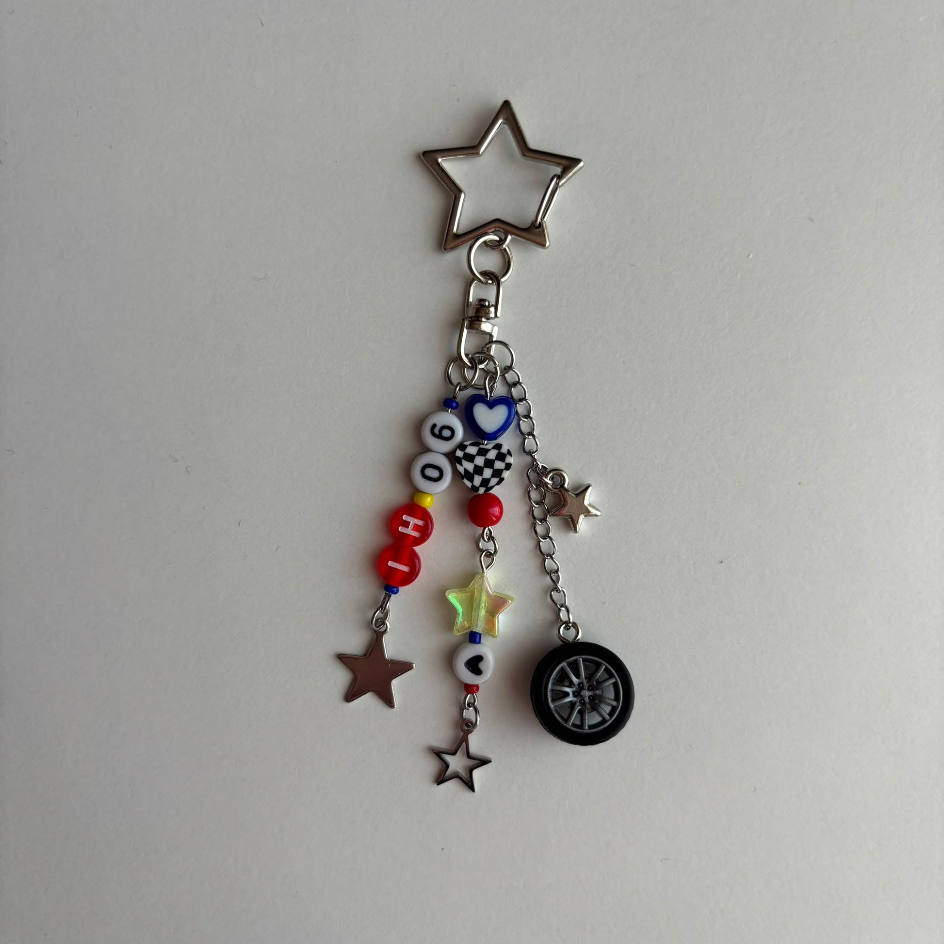 A silver metal keychain charm featuring a star-shaped clasp and dangling charms, including a small tire, star cutouts, checkered pattern, and colorful beads spelling out 'IH06'.