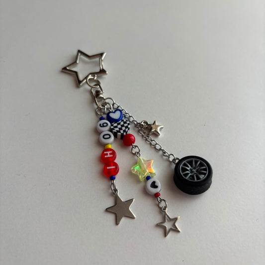 A silver-toned keychain charm featuring a star-shaped keyring and multiple dangling charms, including a black tire, silver stars, a checkered bead, red and white beads, and colorful star and heart beads, shown on a plain light background.