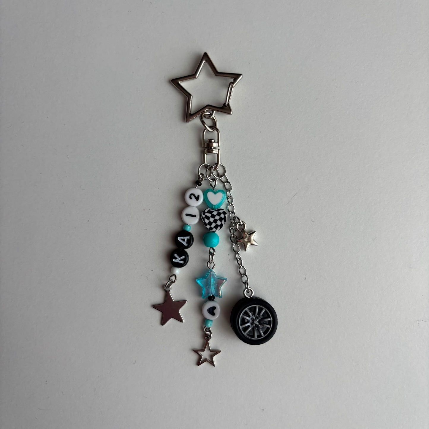A silver-tone keychain charm featuring a star-shaped keyring, a chain with beads, in white, black, and turquoise, a checkered pattern bead, a turquoise star, and a miniature black tire charm, all on a light gray background.