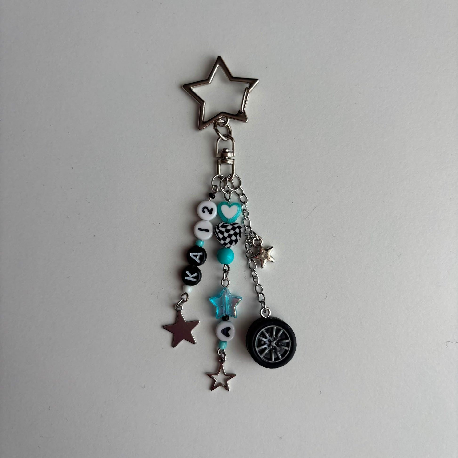 A silver-tone keychain charm featuring a star-shaped keyring, a chain with beads, in white, black, and turquoise, a checkered pattern bead, a turquoise star, and a miniature black tire charm, all on a light gray background.