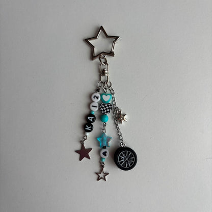 A silver-tone keychain charm featuring a star-shaped keyring, a chain with beads, in white, black, and turquoise, a checkered pattern bead, a turquoise star, and a miniature black tire charm, all on a light gray background.
