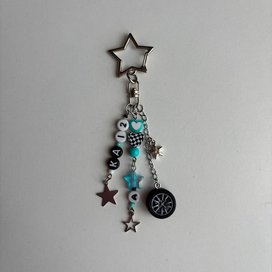 A silver-tone keychain charm featuring a star-shaped keyring, a chain with beads, in white, black, and turquoise, a checkered pattern bead, a turquoise star, and a miniature black tire charm, all on a light gray background.