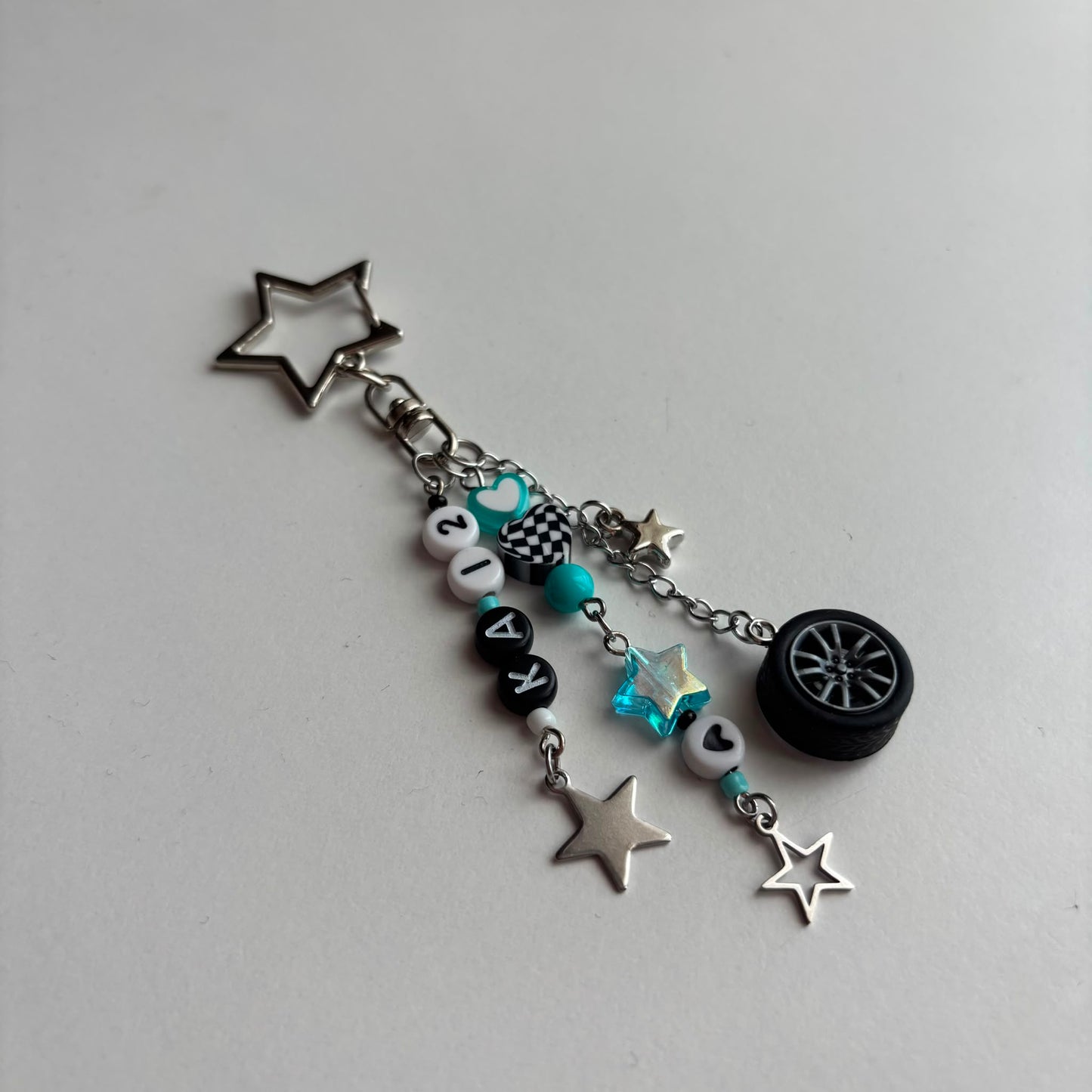 A silver-tone keychain charm featuring a star-shaped keyring, a chain with beads, in white, black, and turquoise, a checkered pattern bead, a turquoise star, and a miniature black tire charm, all on a light gray background.