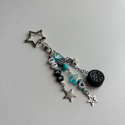 A silver-tone keychain charm featuring a star-shaped keyring, a chain with beads, in white, black, and turquoise, a checkered pattern bead, a turquoise star, and a miniature black tire charm, all on a light gray background.