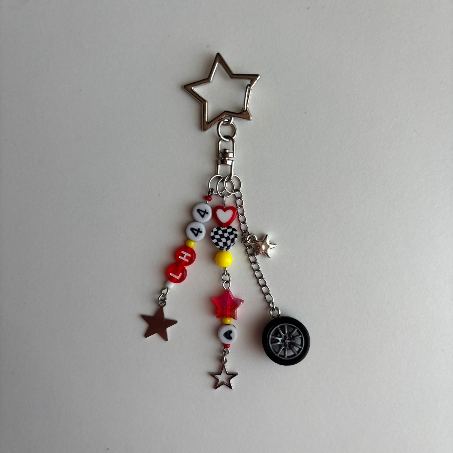 A silver-toned metal keychain charm featuring a star-shaped top, a lobster clasp, and multiple dangling charms including beads spelling 'LH44', small stars, a checkered heart, a red star, and a miniature black tire.