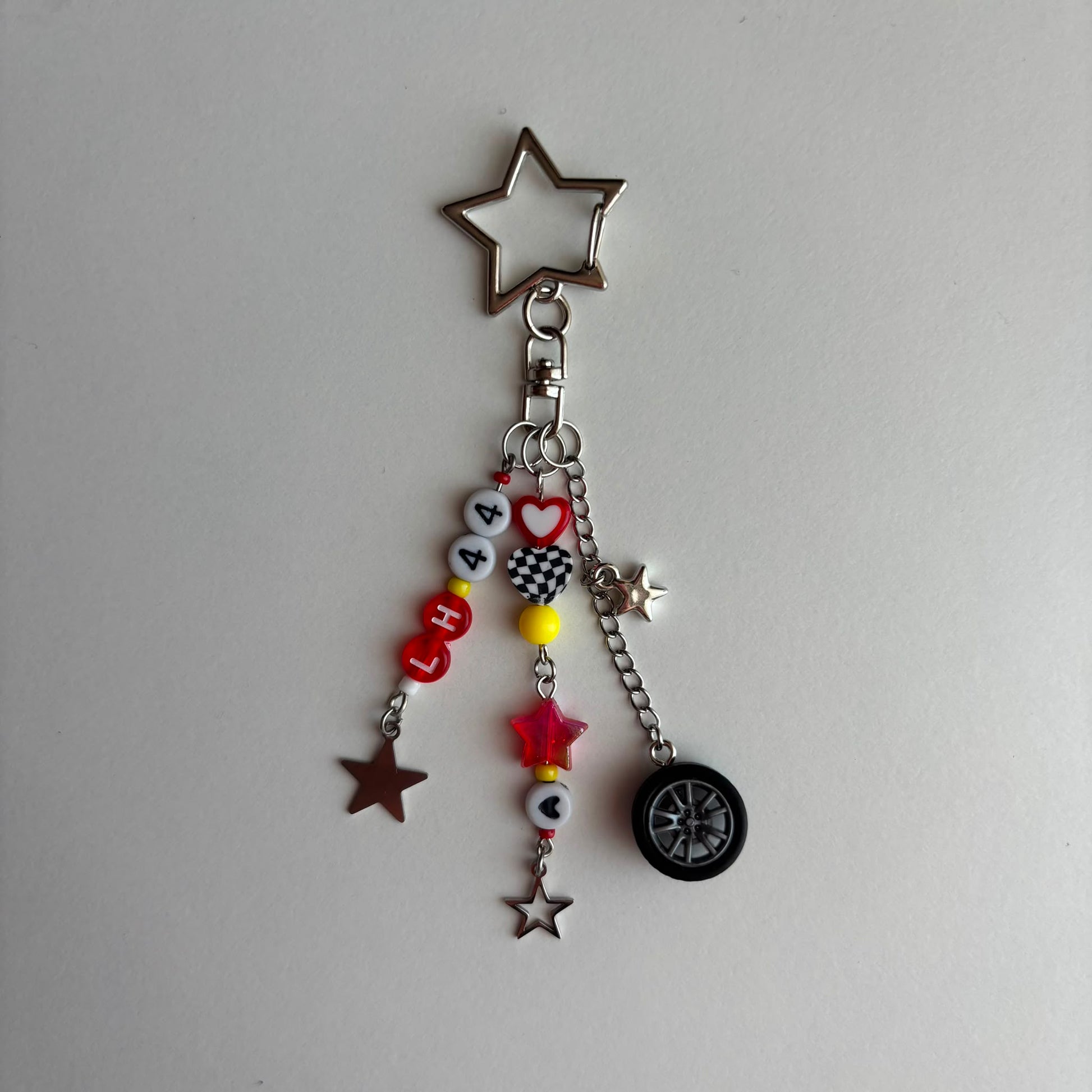 A silver-toned metal keychain charm featuring a star-shaped top, a lobster clasp, and multiple dangling charms including beads spelling 'LH44', small stars, a checkered heart, a red star, and a miniature black tire.