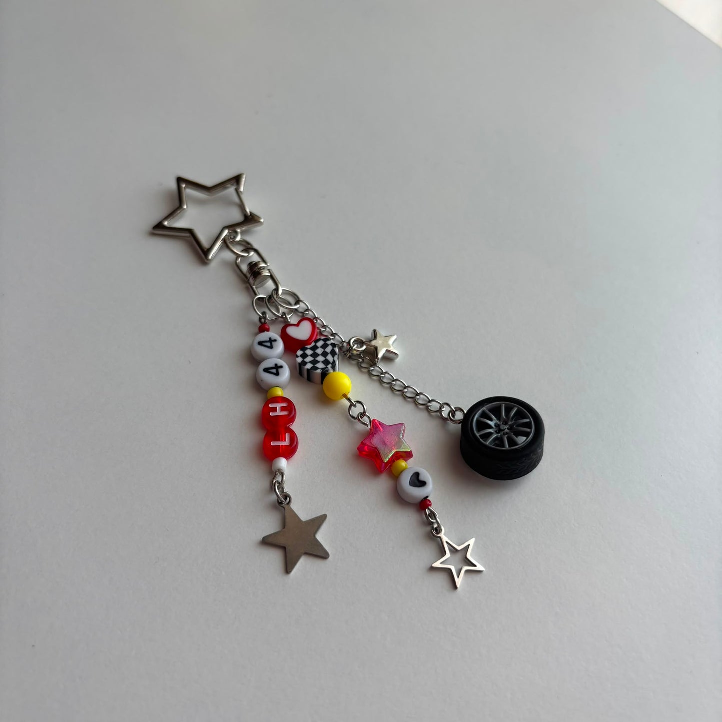 A silver-toned keychain charm featuring a star-shaped ring, a small black tire charm, and multiple dangling charms including star cutouts, a checkered piece, red and white letter beads spelling 'LH44', and a small yellow bead, photographed on a plain light gray surface.