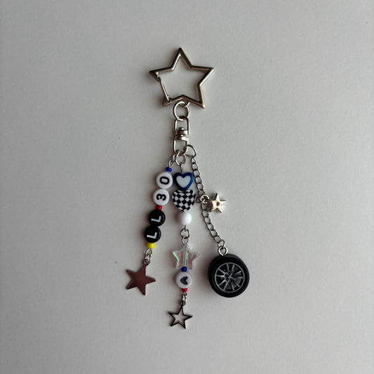 A silver metal keychain charm featuring a star-shaped ring at the top and dangling charms including black and white beads spelling 'LL30', star charms, a checkered heart, and a small black tire charm, photographed on a plain light gray background.