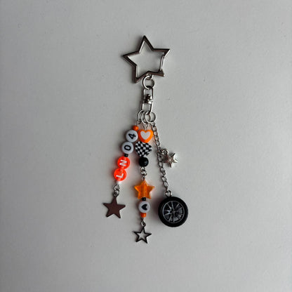 A silver metal keychain charm featuring a large star-shaped ring at the top and multiple dangling charms including orange and white beads spelling 'LN04', small stars, a checkered flag motif, and a miniature tire.