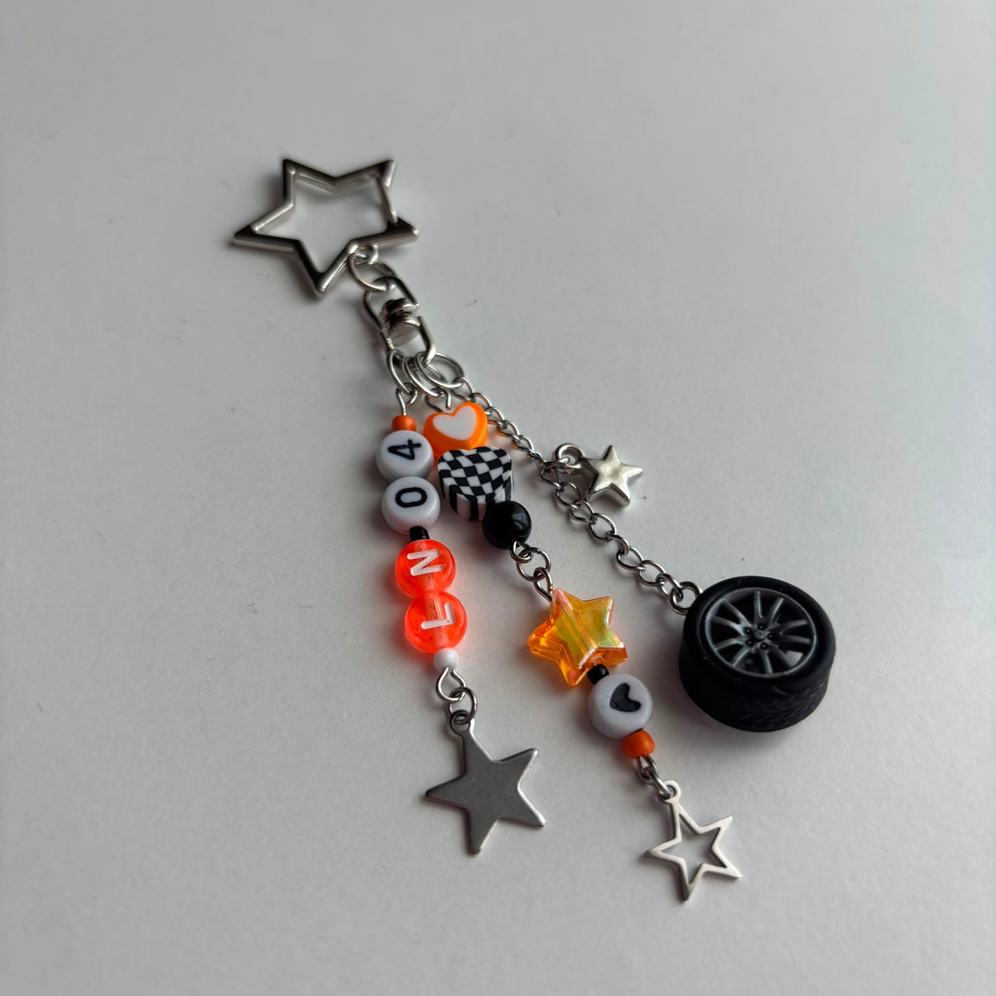 A silver-toned metal keychain charm featuring a star-shaped top, a checkered flag motif, white and orange letter beads spelling 'LN04', a small black tire charm, and multiple dangling star charms on a plain light gray background.