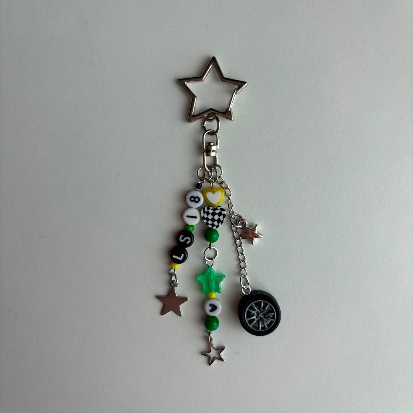 A silver metal keychain charm featuring a star-shaped ring at the top, a lobster clasp, and multiple dangling charms including beads spelling 'LS18', a small heart, a star, a mini tire, and green and silver star accents, shown on a plain light gray background.