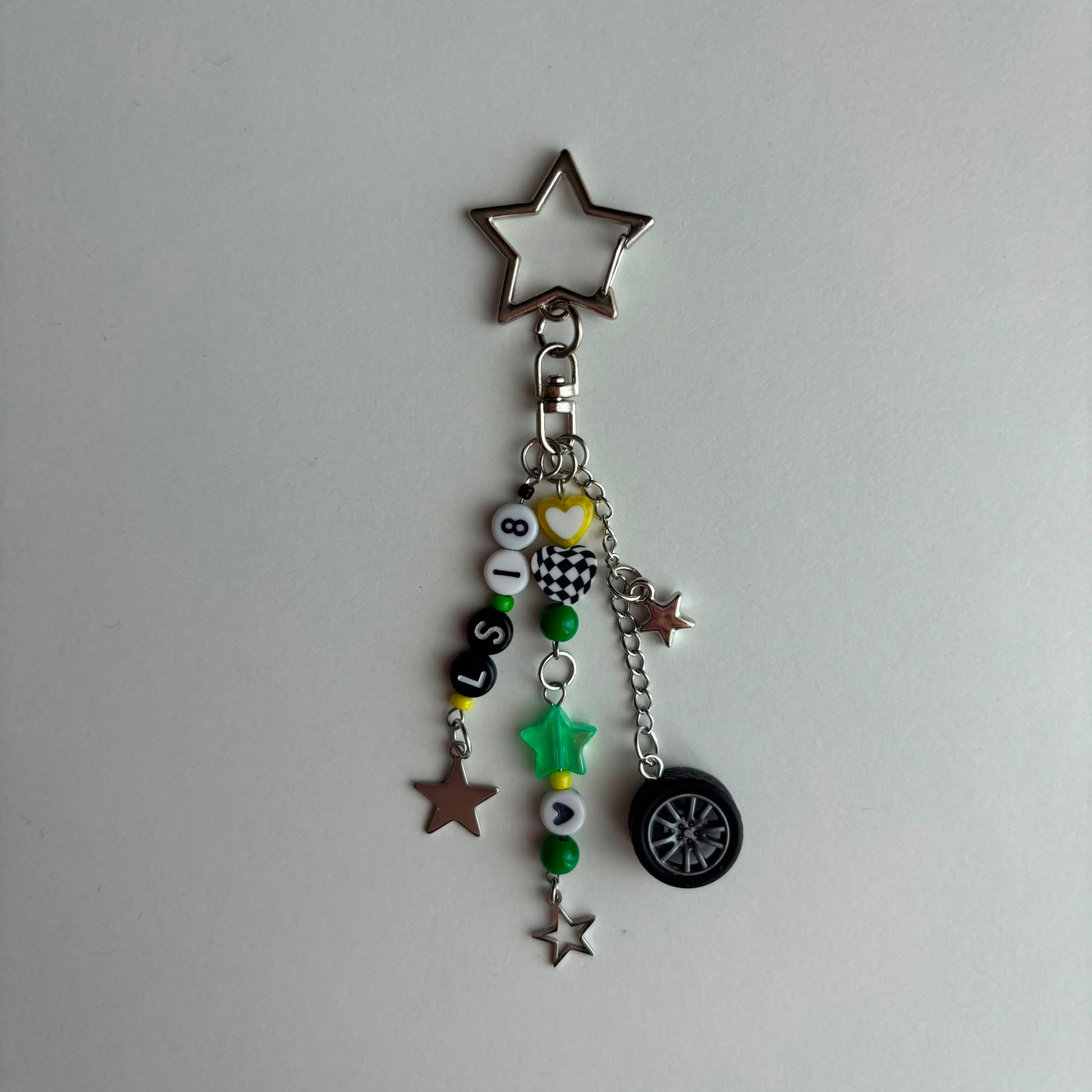 A silver metal keychain charm featuring a star-shaped ring at the top, a lobster clasp, and multiple dangling charms including beads spelling 'LS18', a small heart, a star, a mini tire, and green and silver star accents, shown on a plain light gray background.