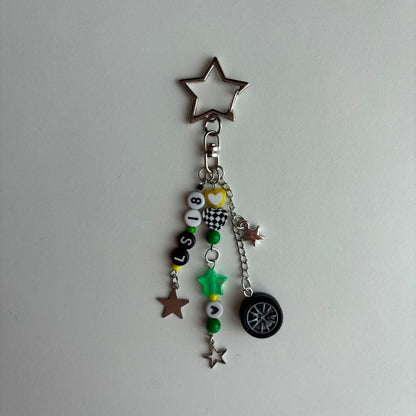 A silver metal keychain charm featuring a star-shaped ring at the top, a lobster clasp, and multiple dangling charms including beads spelling 'LS18', a small heart, a star, a mini tire, and green and silver star accents, shown on a plain light gray background.
