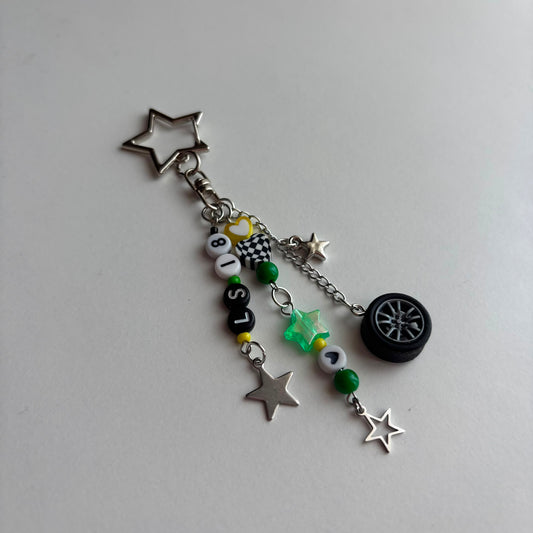 A silver-toned keychain charm featuring a star-shaped keyring, dangling chain, beads spelling 'LS18', green and white beads, a small tire charm, and multiple silver star charms on a light gray background.