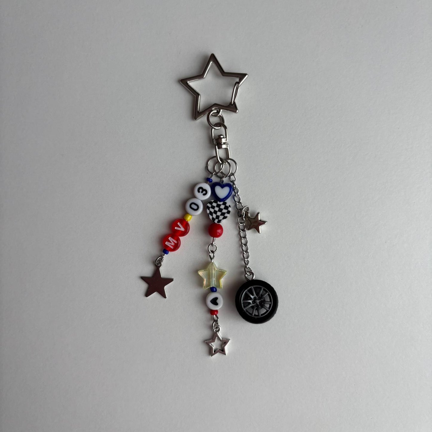 A silver metal keychain charm featuring a star-shaped clasp and multiple dangling charms, including red and white beads, a small heart-shaped charm, a mini tire, and star charms, shown against a plain light gray background.