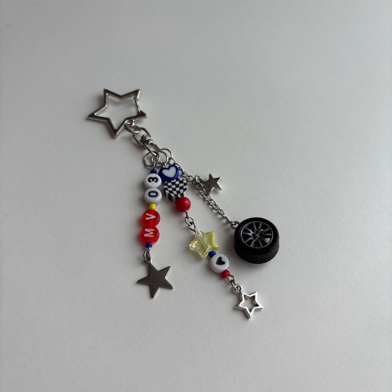 A silver-toned keychain charm featuring a star-shaped ring, a miniature black car wheel, a checkered flag, and red, white, and blue beads spelling 'MV03' on a plain light gray background.