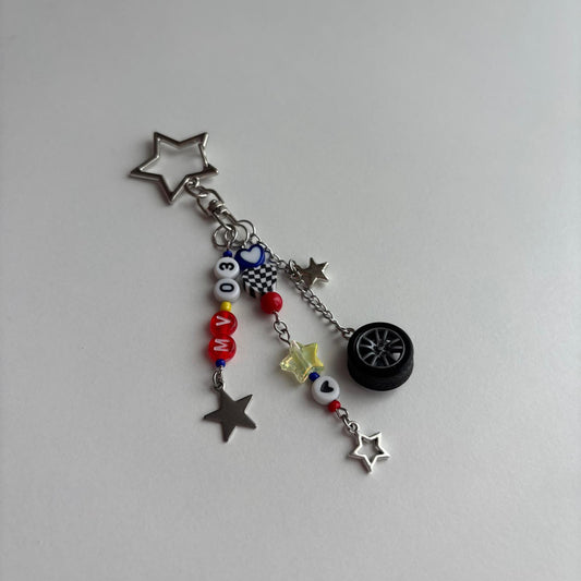 A silver-toned keychain charm featuring a star-shaped ring, a miniature black car wheel, a checkered flag, and red, white, and blue beads spelling 'MV03' on a plain light gray background.