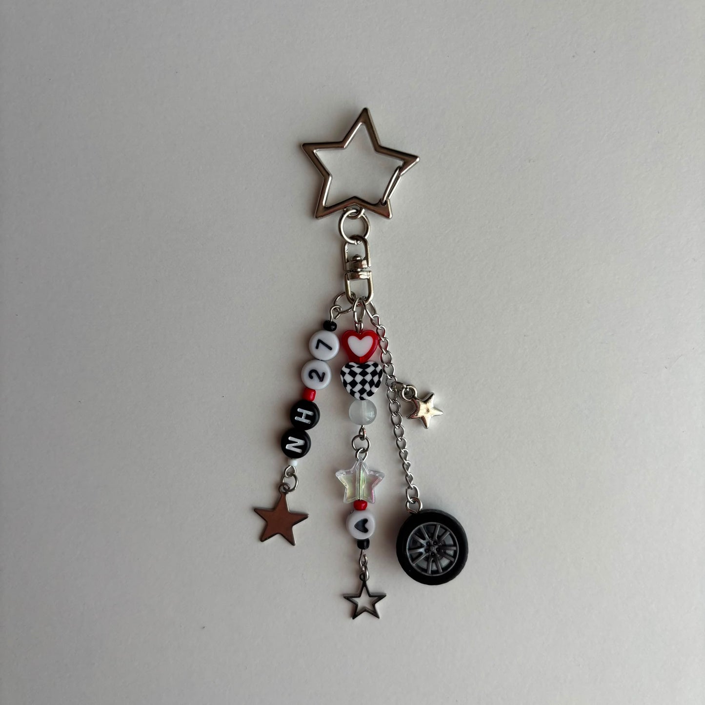 A silver-toned keychain charm featuring a large star-shaped top, a clasp, and dangling charms including black and white beads spelling '27', a red heart, a checkered flag motif, a small white star, a brown star, and a black tire charm, all photographed on a plain light background.