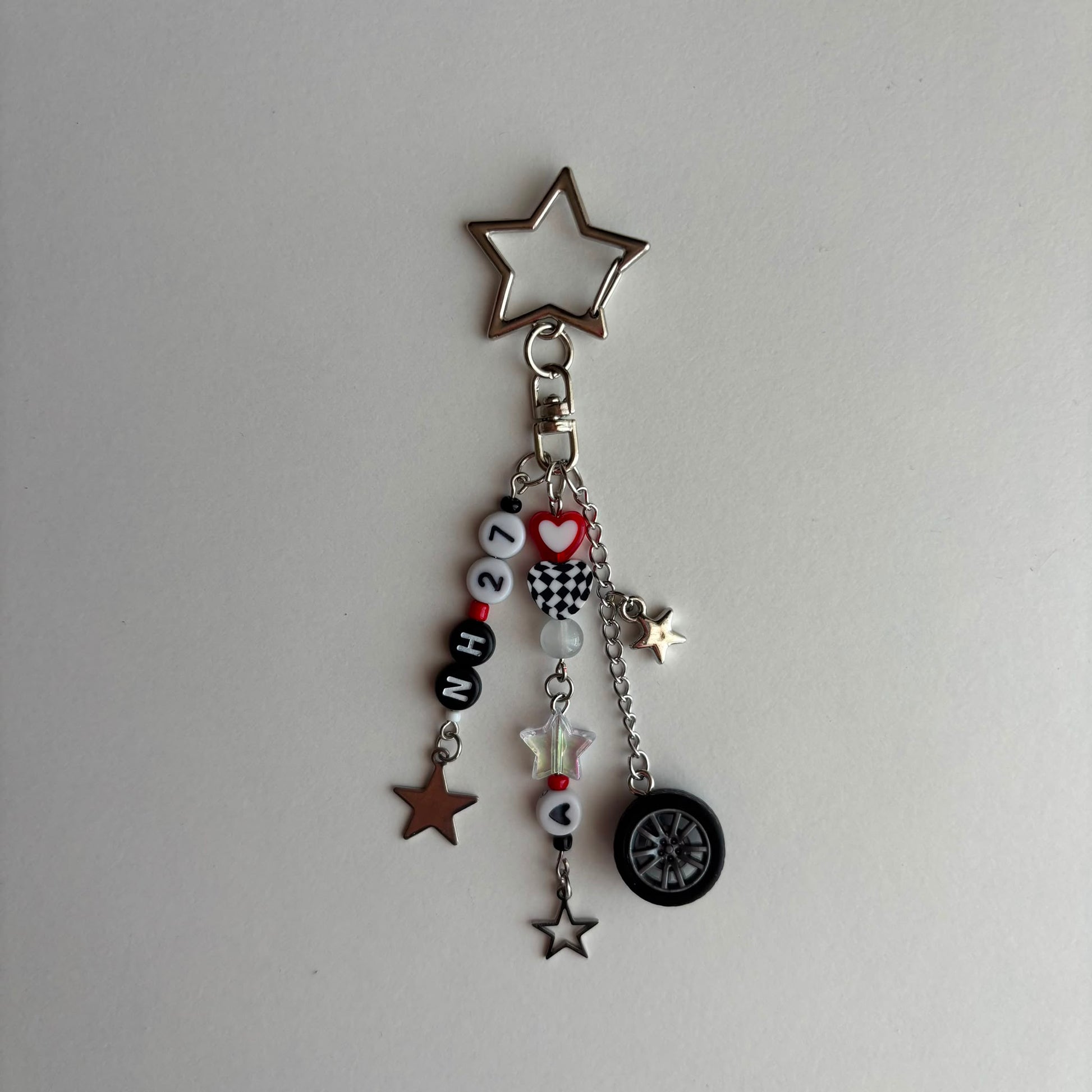 A silver-toned keychain charm featuring a large star-shaped top, a clasp, and dangling charms including black and white beads spelling '27', a red heart, a checkered flag motif, a small white star, a brown star, and a black tire charm, all photographed on a plain light background.